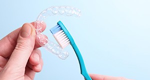 Hands holding blue toothbrush close to clear aligner with pale blue background