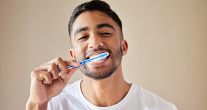 Man in white t-shirt brushing teeth with tan background